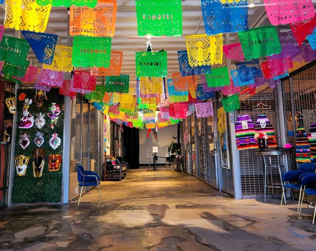 Colorful papel picado banners hanging across an indoor market walkway, with small shops, chairs, and handcrafted items displayed on both sides.
