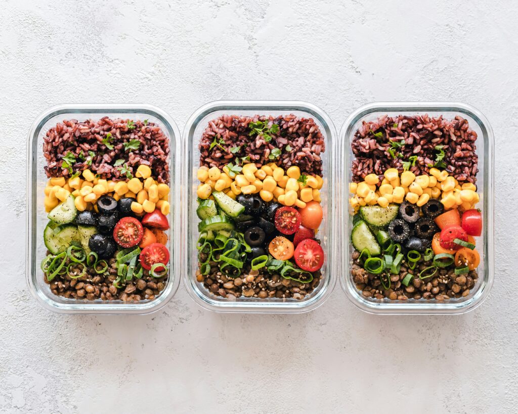 Three glass meal-prep containers filled with layered grains, corn, cucumbers, olives, tomatoes, lentils, and herbs, arranged side by side.