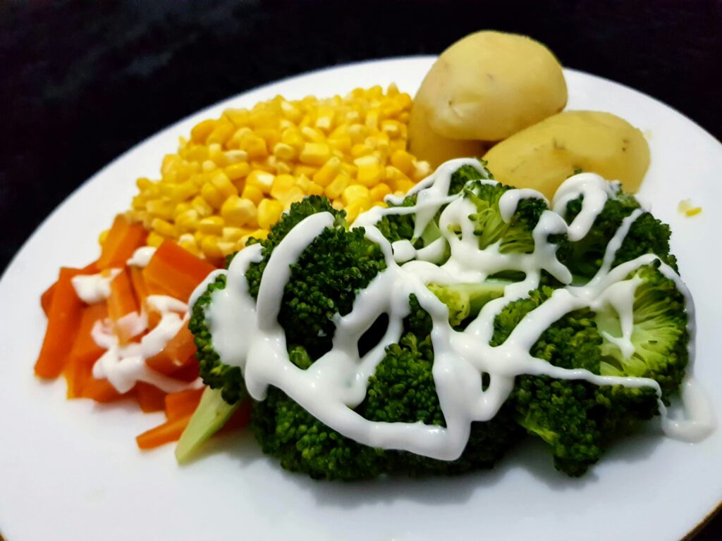 Plate of steamed broccoli drizzled with white sauce, served with corn kernels, boiled potatoes, and sliced carrots.