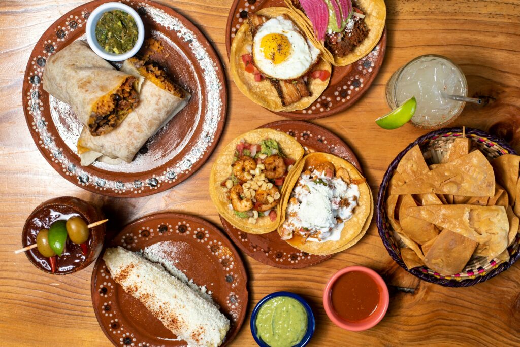 Assorted Mexican dishes including tacos, tortillas, salsas, and side dishes arranged on a wooden table, viewed from above.