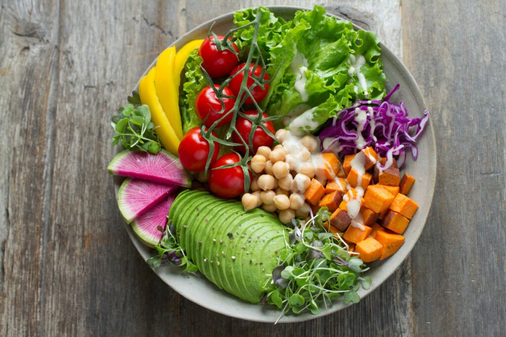 Colorful vegetable bowl with sliced avocado, cherry tomatoes, chickpeas, sweet potatoes, leafy greens, and microgreens arranged on a plate.
