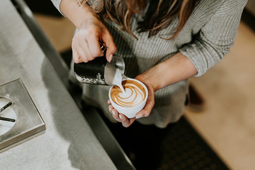 Barista pouring steamed milk into a cup of coffee to create latte art, viewed from above at a café counter.