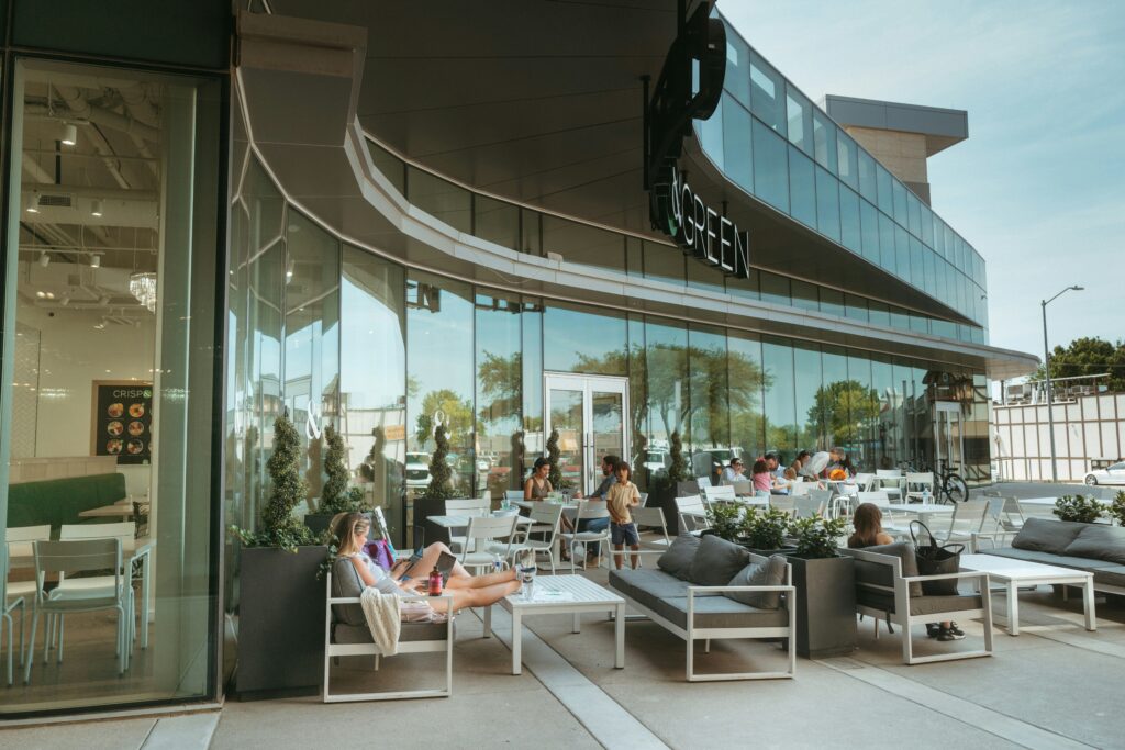 People seated at outdoor tables on a modern restaurant patio with glass walls, white furniture, and a curved facade in a shopping district.