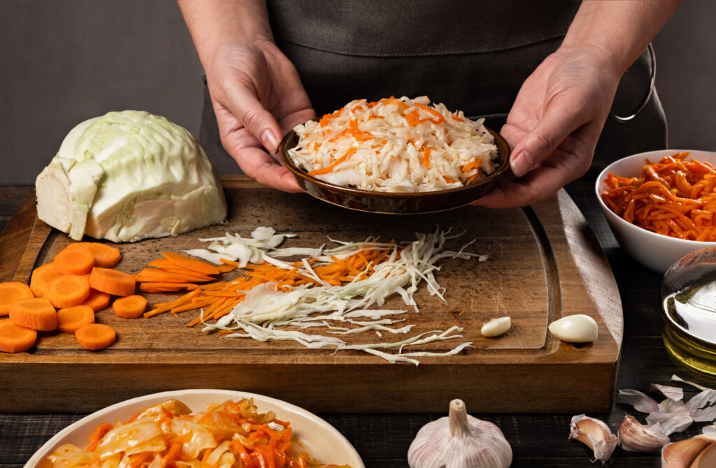 Hands holding a bowl of curtido slaw over a cutting board with sliced cabbage, carrots, garlic, and olive oil used in preparation.