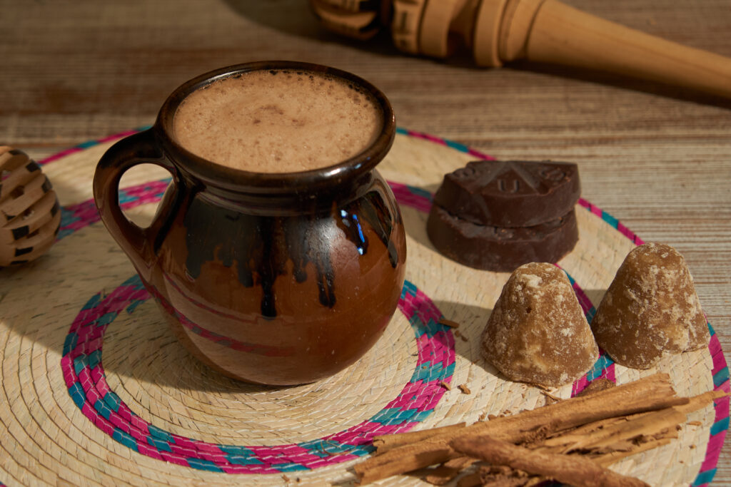 Clay mug of traditional Mexican hot chocolate on a woven mat, surrounded by cinnamon sticks, piloncillo cones, chocolate, and a wooden molinillo.