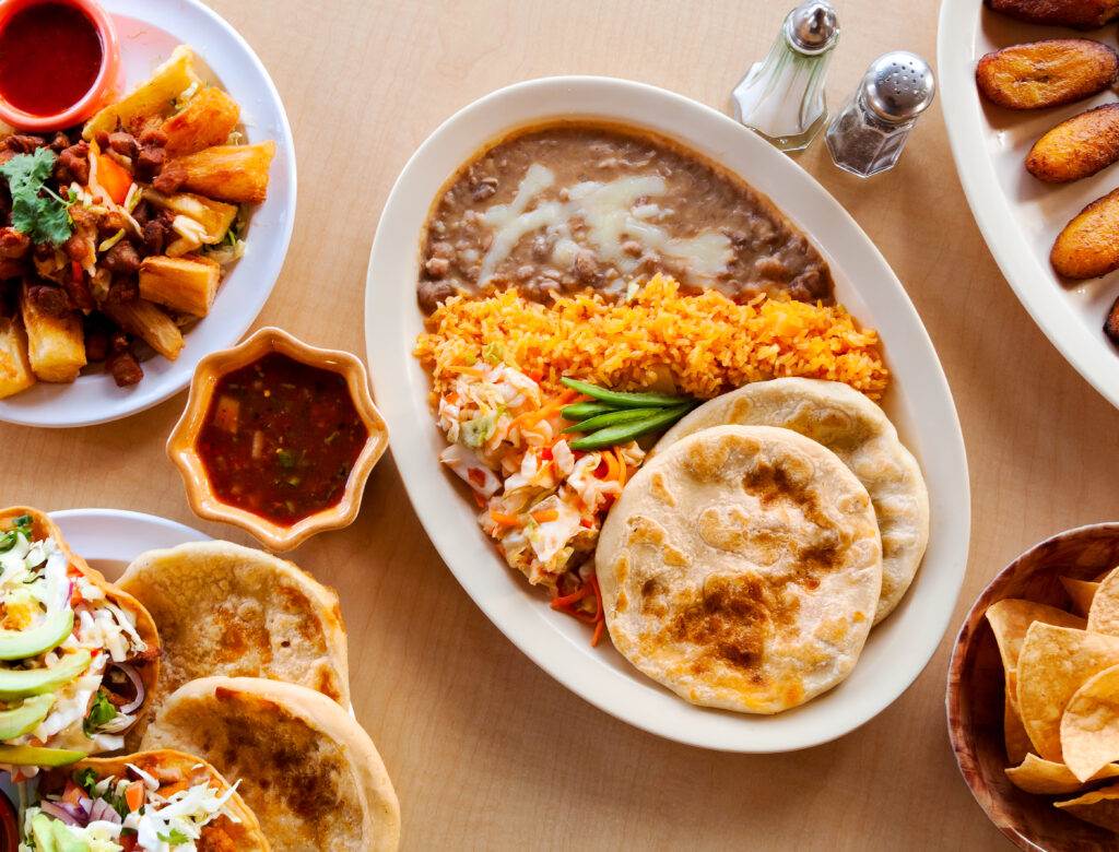 Platter of traditional Salvadoran food with pupusas, rice, refried beans, curtido slaw, and green beans, surrounded by chips, fried plantains, and salsa.