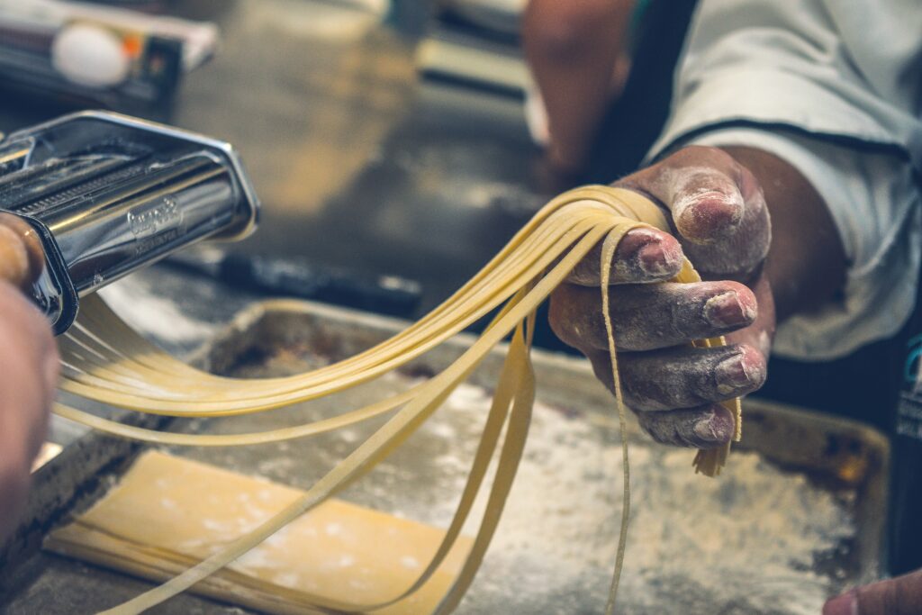 Hands dusted with flour pulling fresh pasta strands from a pasta machine over a floured work surface.