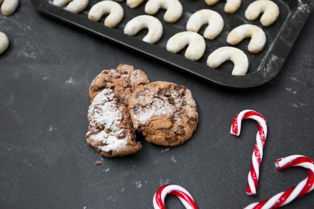 Powdered sugar–dusted cookies on a dark surface, with a candy cane and additional cookies blurred in the background.