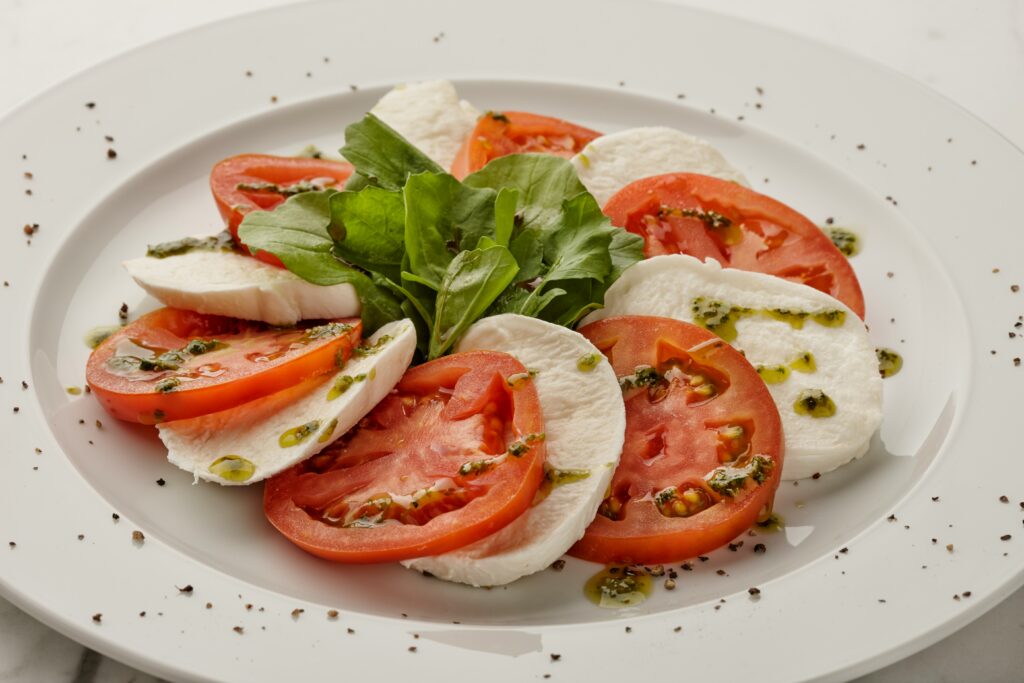 Caprese salad with sliced tomatoes, fresh mozzarella, basil leaves, and olive oil drizzle on a white plate.