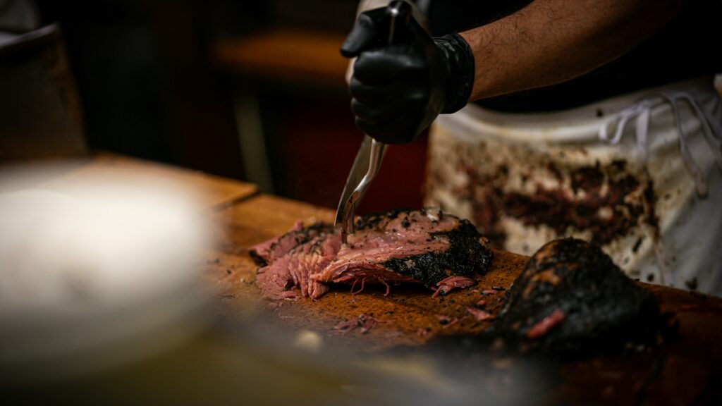 Cook wearing black gloves slicing smoked brisket on a wooden cutting board, showing the pink interior and charred crust.