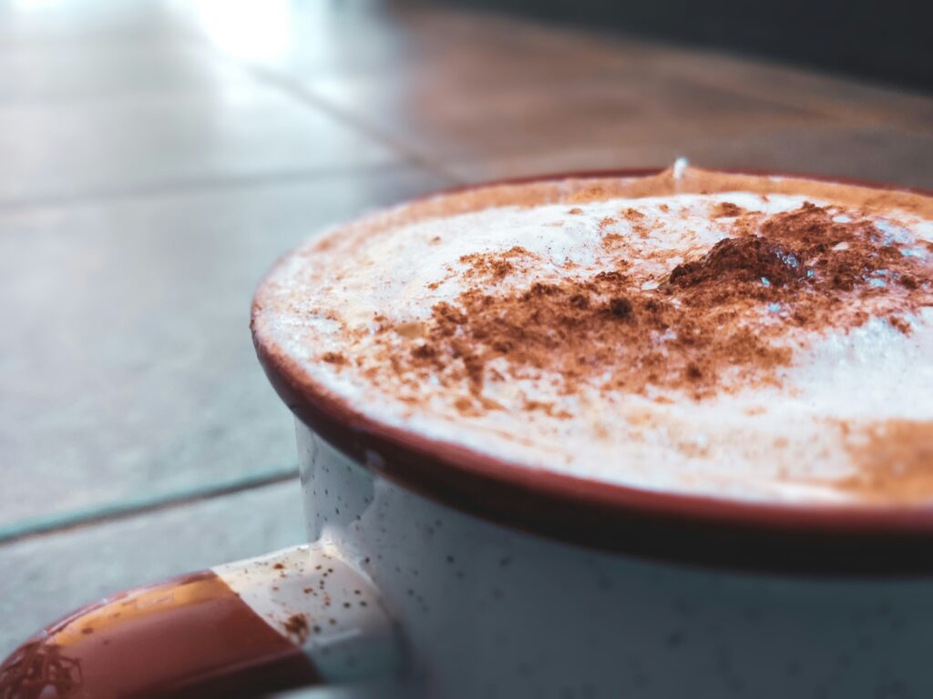 Close-up of a ceramic mug filled with a frothy hot drink, topped with foam and sprinkled cinnamon, resting on a tiled surface.