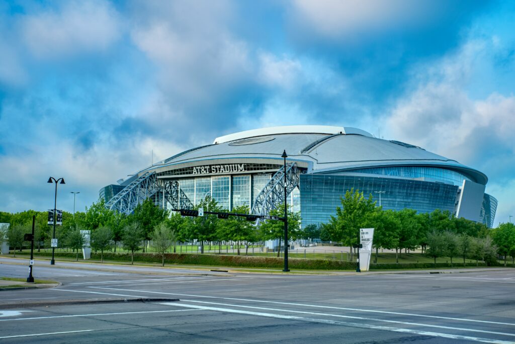 Modern stadium exterior with curved glass and metal facade, viewed from a street intersection under a partly cloudy blue sky.
